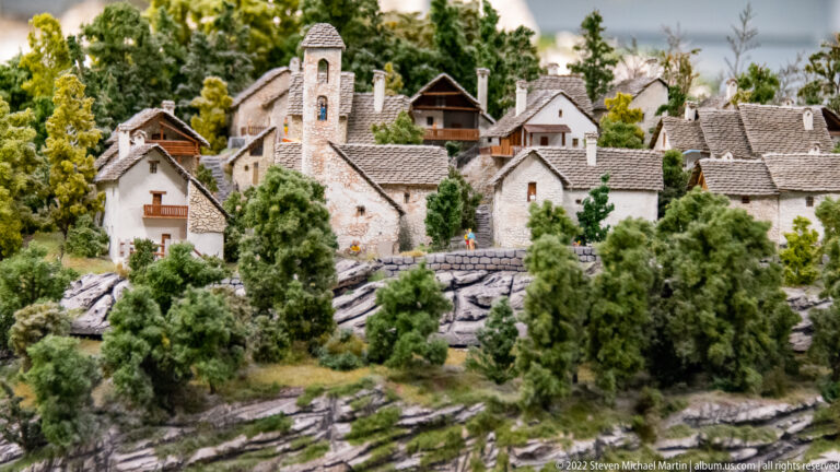 Miniature alpine village with gray-roofed houses, a stone church tower, trees, and rocky slopes.