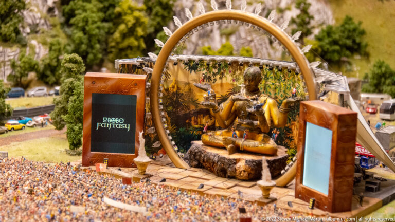 Fantasy concert stage with a large circular arch, golden seated figure, screens, and a dense miniature crowd.