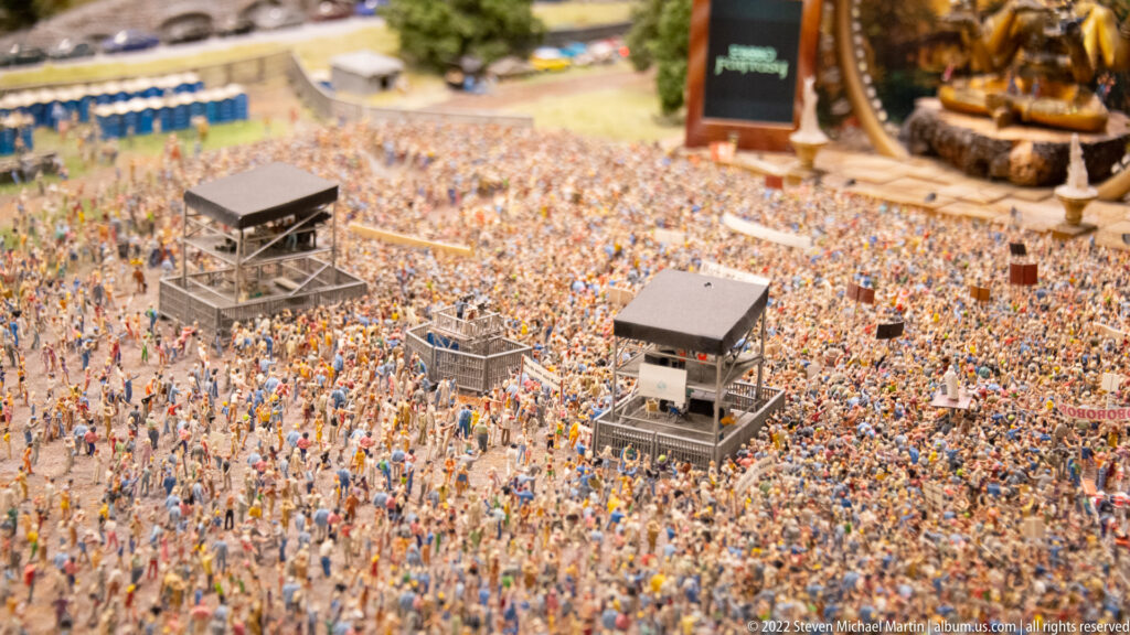 Dense miniature festival crowd surrounding two elevated control towers, with the concert stage partly visible beyond.