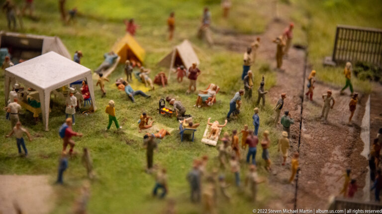 Miniature campground with tents, a canopy, people relaxing on grass, and a gravel path.