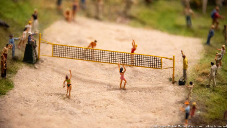 Miniature volleyball players on a sand court with a net and spectators standing along the edges.