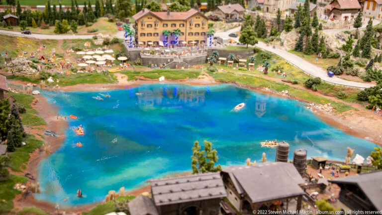 Bright blue miniature lake with swimmers, boats, lakeside buildings, roads, and forested mountain slopes.