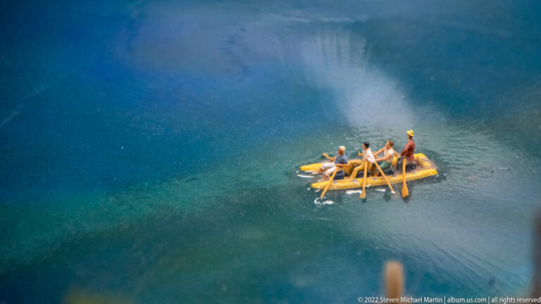 Yellow miniature raft with several paddlers crossing blue water, surrounded by faint ripples.