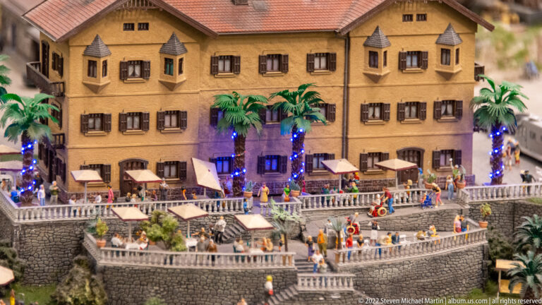Ochre miniature resort building with palm trees, illuminated terraces, tables, and many diners.