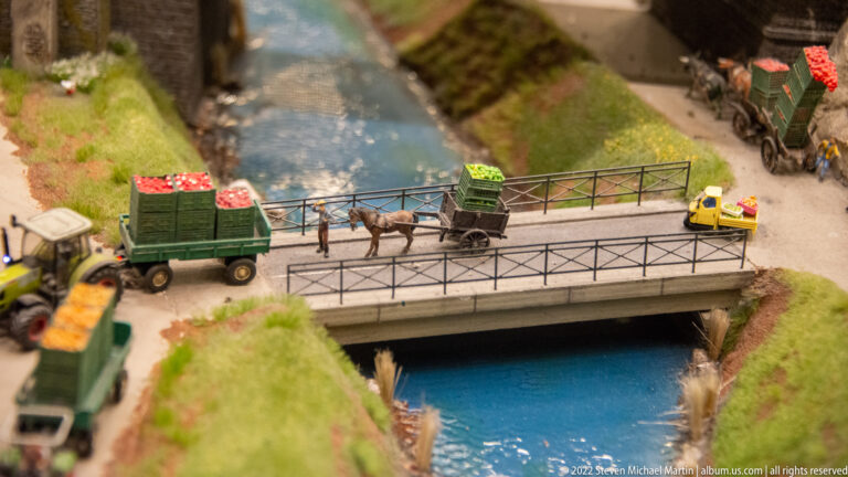 Horse-drawn cart, trucks, and small vehicles crossing a miniature bridge over blue water.