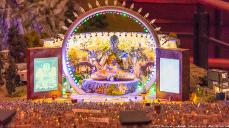 Illuminated fantasy concert stage with circular arch, central seated figure, side screens, and glowing audience lights.