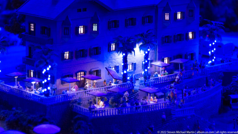 Blue-lit miniature resort building at night with glowing windows, palm trees, and occupied terraces.