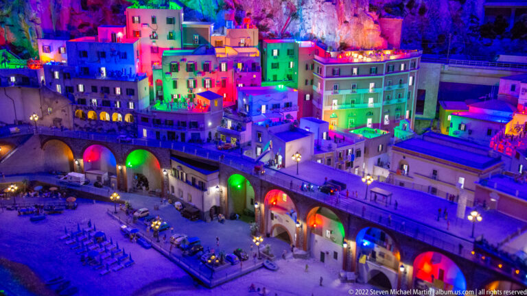 Colorfully lit miniature city above a waterfront promenade with large arches and outdoor seating.