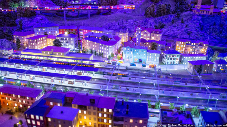 Nighttime miniature town with a multi-track railway station, platforms, trains, and illuminated buildings.