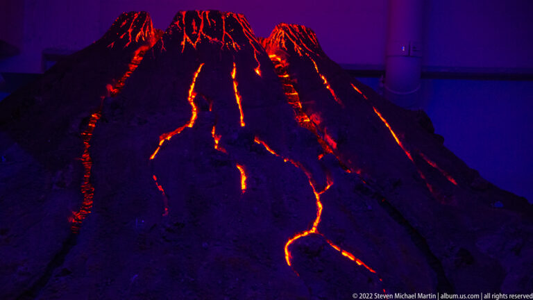 Dark miniature volcano with bright orange-red lava streams glowing down its slopes at night.