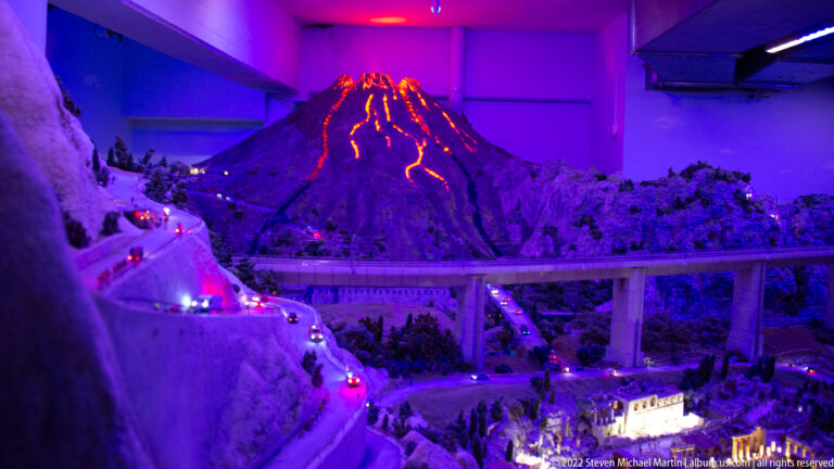 Glowing miniature volcano above a winding road, vehicles, rocky terrain, and an elevated viaduct.