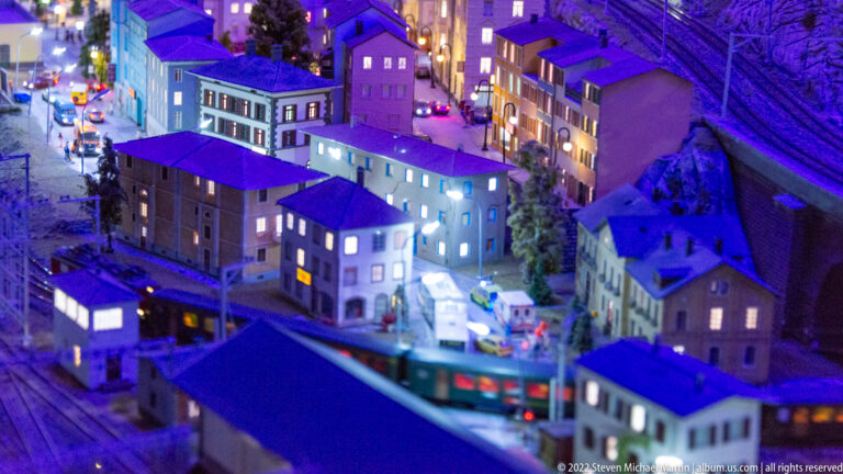 Miniature nighttime neighborhood beside railway tracks, with illuminated buildings and a passing train in front.
