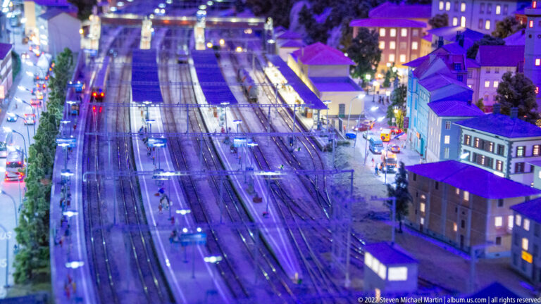 Nighttime miniature rail yard with many parallel tracks, platforms, lights, and buildings along the right edge.