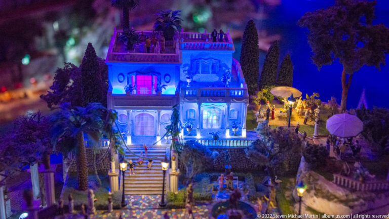 Blue-lit miniature villa with terraces, stairs, trees, umbrellas, and groups of people in the garden.
