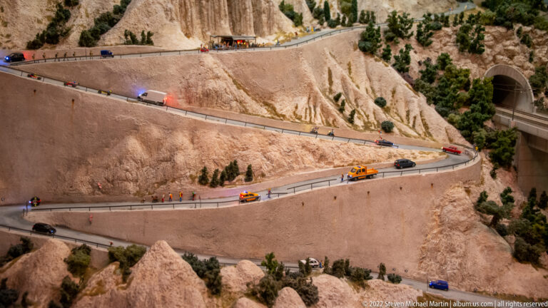 Miniature mountain road with hairpin turns, cars, trucks, rocky slopes, sparse trees, and tunnel portals.
