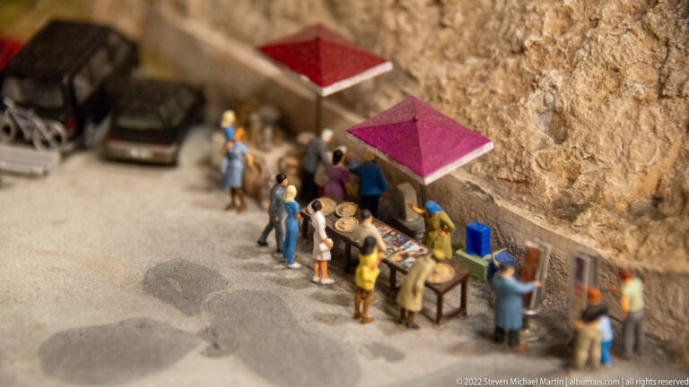 Miniature shoppers and vendors at small market tables with red awnings beside a rock wall.