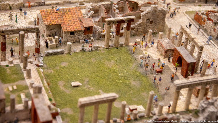 Miniature stone ruins with columns, roofless walls, a grassy courtyard, and numerous visitors.