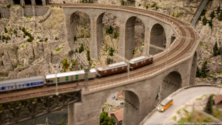 Passenger train crossing a curved stone-arch viaduct above a rocky mountain road.