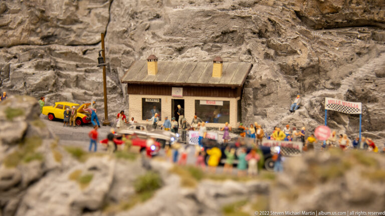 Small miniature station building against a rock face, with crowds, vehicles, barriers, and a tunnel nearby.
