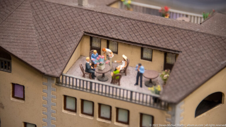 Miniature people gathered around tables on a recessed rooftop terrace within a pitched-roof building.