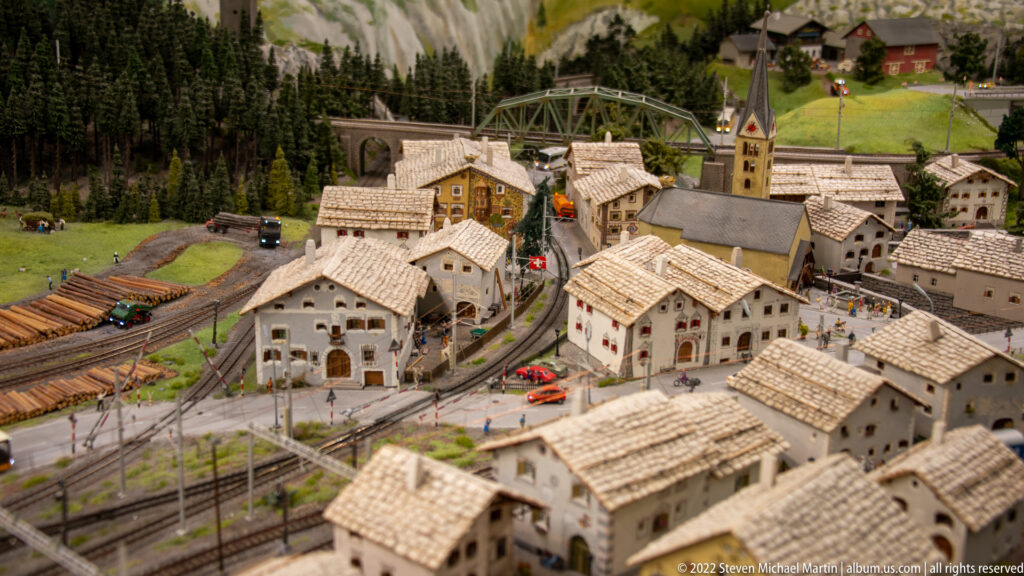 Swiss-themed miniature alpine village with church, railway tracks, pale-roofed houses, roads, and red Swiss flags.