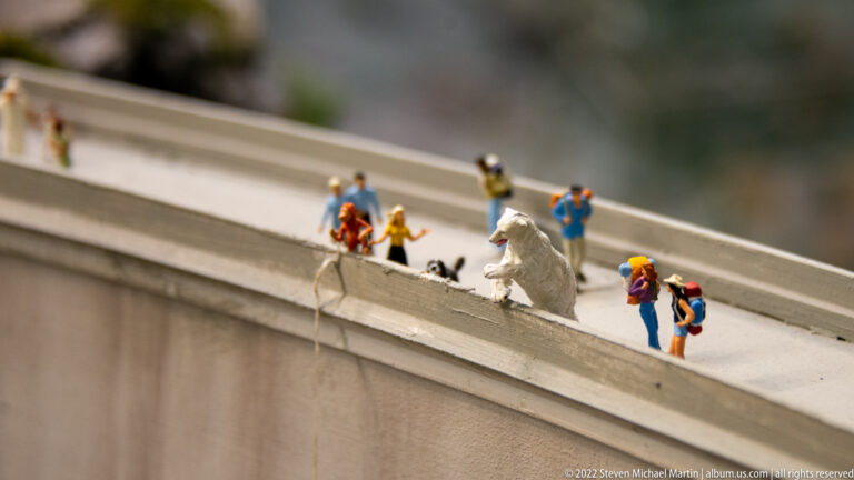 Miniature climbers or rescuers on a narrow elevated edge, with one figure suspended by a rope near a white form.