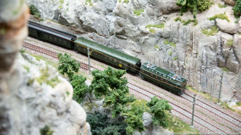 Green electric locomotive pulling passenger cars through a rocky miniature railway cutting with overhead wires.