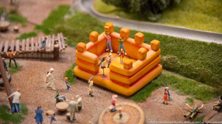 Orange miniature inflatable play structure on grass, with children, adults, tables, and a gravel recreation area.