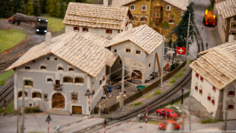 Swiss miniature village with white alpine houses, railway tracks and catenary, road traffic, and a red Swiss flag.