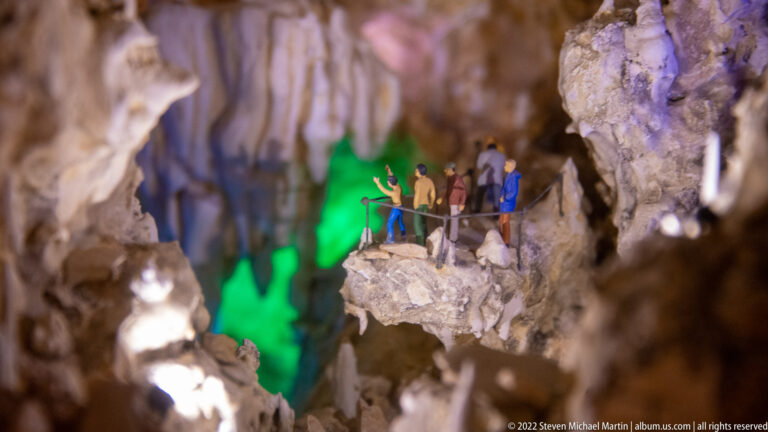 Miniature visitors stand behind a railing on a rocky cave ledge, with vivid green light illuminating the cavern behind them.