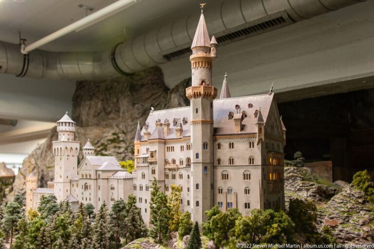 Miniature Neuschwanstein Castle stands among model trees and rocks beneath visible exhibition ductwork.