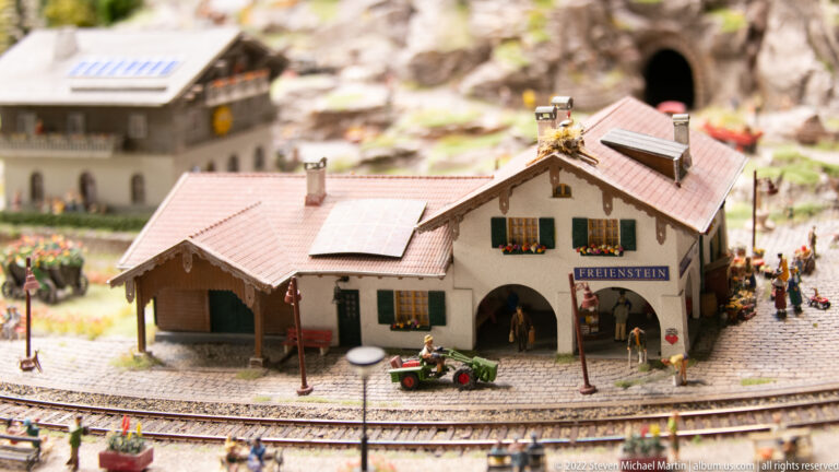 Small alpine station building with pitched roof, flower boxes, passengers, and railway tracks in the foreground.