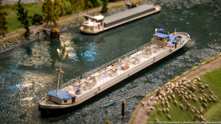 Long model barge and smaller boat travel along a canal beside a flock of miniature sheep.