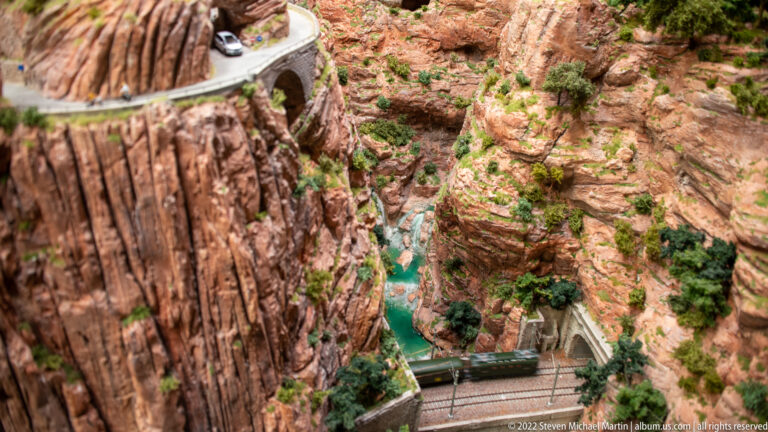 Miniature train and bridge crossing a steep red-rock canyon above turquoise water.