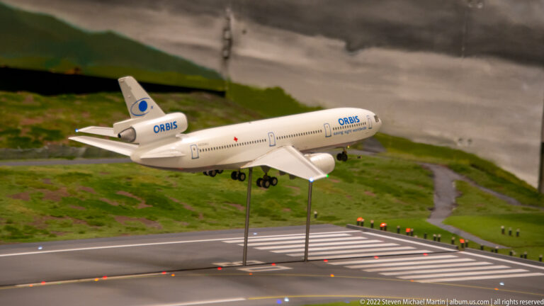 ORBIS Flying Eye Hospital model aircraft descends above a miniature runway, supported visibly by thin display rods.