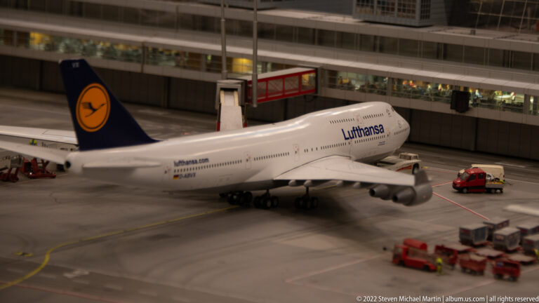 Lufthansa Boeing 747 model parked beside a miniature terminal with ground-service vehicles and baggage equipment.