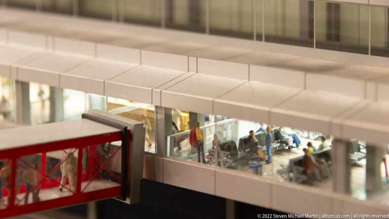 Miniature airport passengers gather inside a glass-walled terminal concourse beside a red enclosed connector.