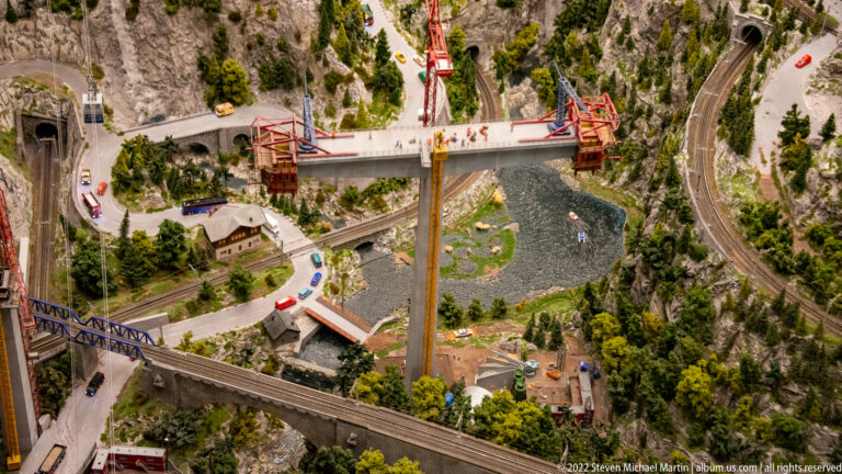Overhead view of a miniature bridge construction site with cranes, river, roads, tunnels, and rail viaduct.