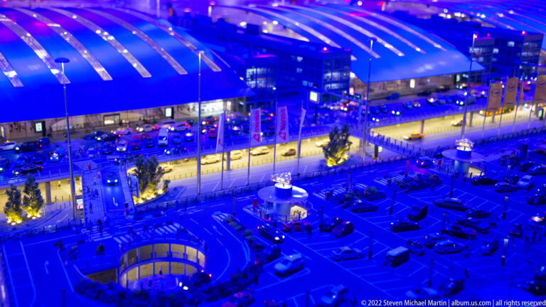 Blue-lit miniature airport terminal complex with roads, parking areas, traffic, and illuminated interiors.