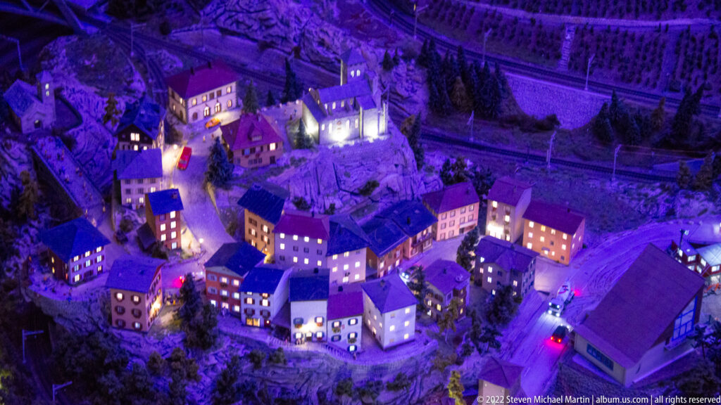 Illuminated miniature alpine village clustered on a wooded slope beneath railway tracks at night.