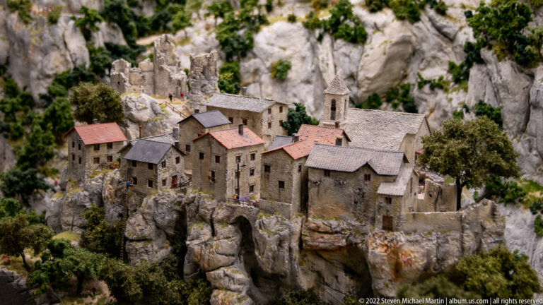 Miniature stone village with church tower perched among steep gray limestone cliffs and trees.
