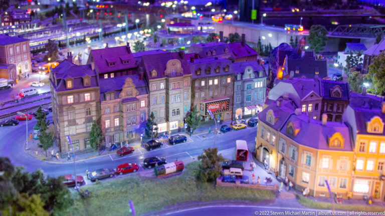 Miniature European townhouses line a curved street with cars, pedestrians, and illuminated windows at night.