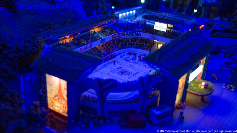 Open-roof miniature ice hockey arena with players, spectators, rink boards, and illuminated screens under blue light.