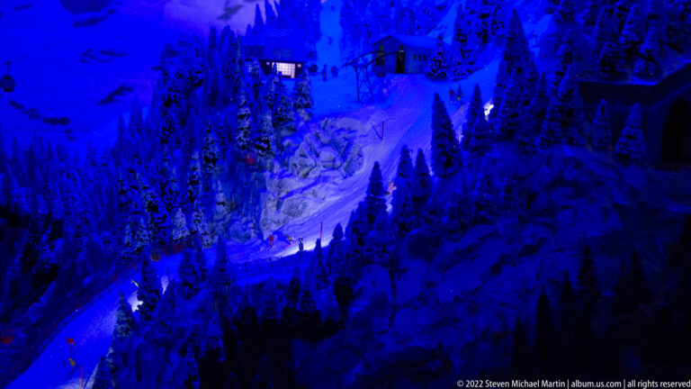 Snowy miniature alpine landscape with conifers, rocky slopes, small building, and tunnel portal under blue night lighting.