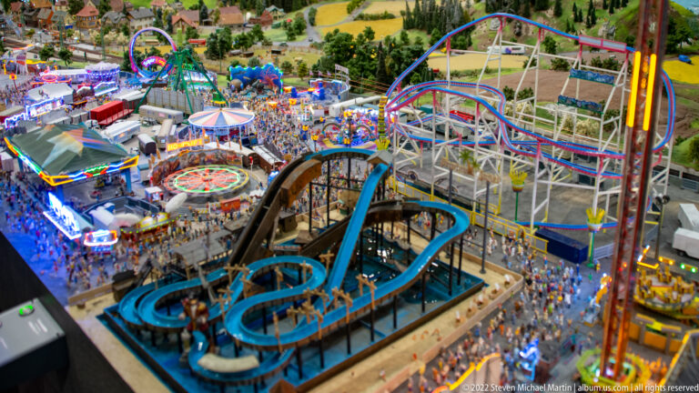 Miniature fairground with blue water ride, roller-coaster tracks, crowds, stalls, and colorful amusement structures.