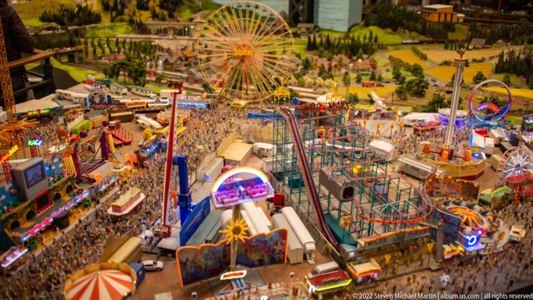 Large miniature fairground packed with a Ferris wheel, roller coaster, thrill rides, tents, and crowds.