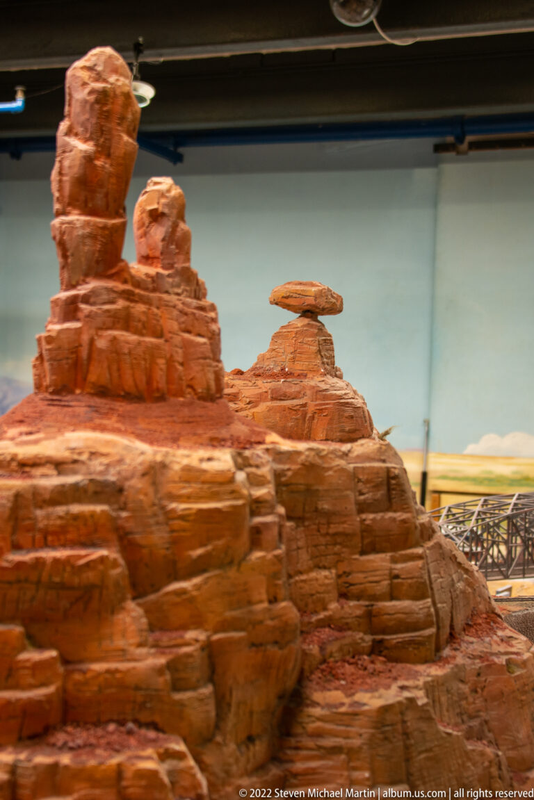 Miniature red-rock desert with layered sandstone buttes, balanced formations, and distant bridge.