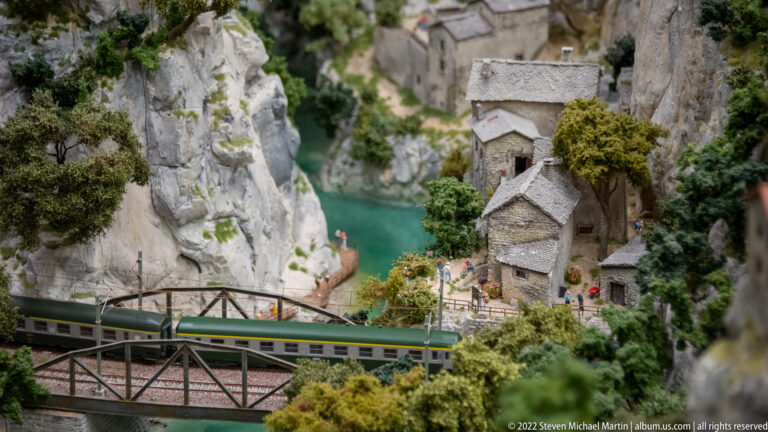 Passenger train crossing a steel truss bridge in front of a miniature gorge village and turquoise water.