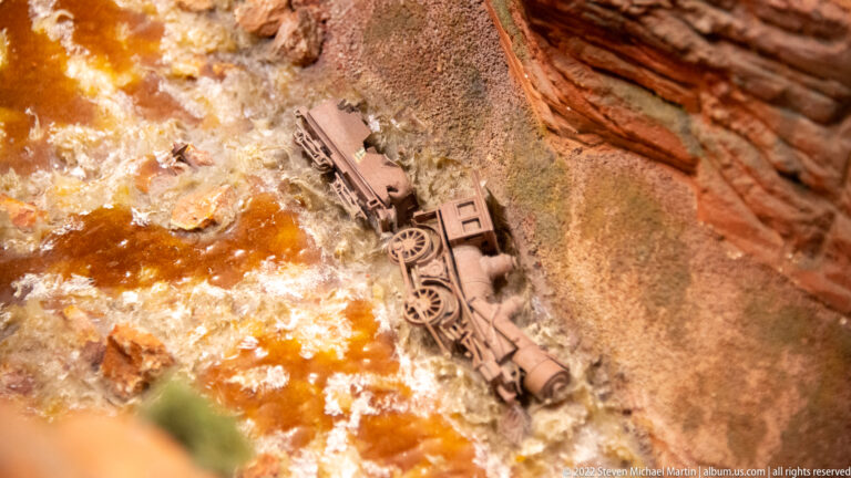 Overturned miniature steam locomotive lies in foaming water at the base of a steep red-rock canyon.