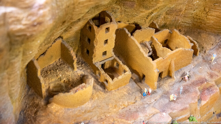 Miniature Ancestral Puebloan-style cliff dwelling with roofless rooms built beneath a broad rock overhang.